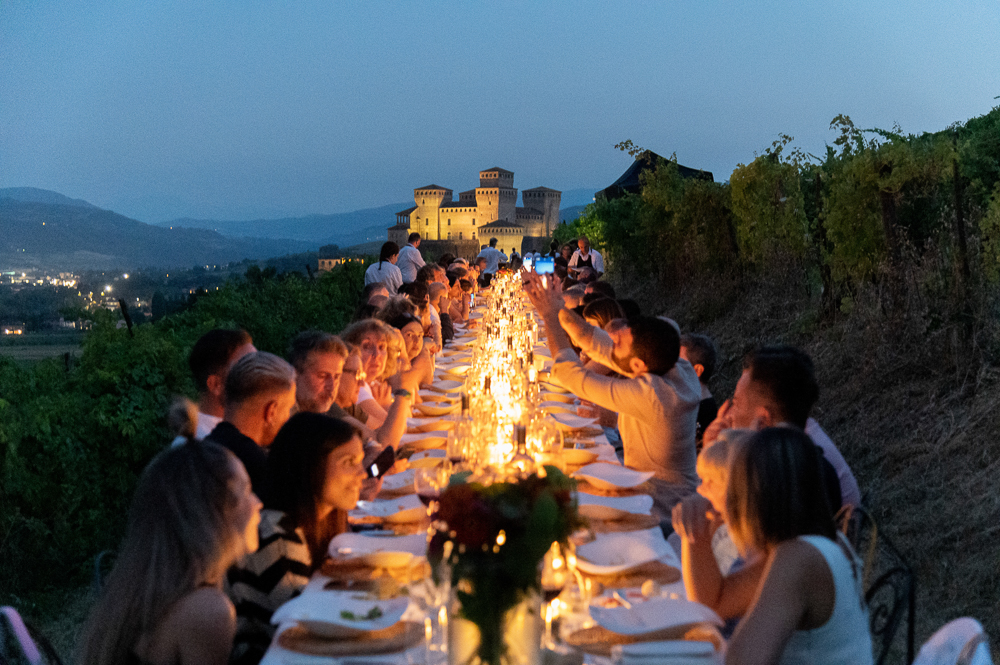 Cena in vigna al tramonto con lungo tavolo illuminato da candele, numerosi commensali e castello sullo sfondo.